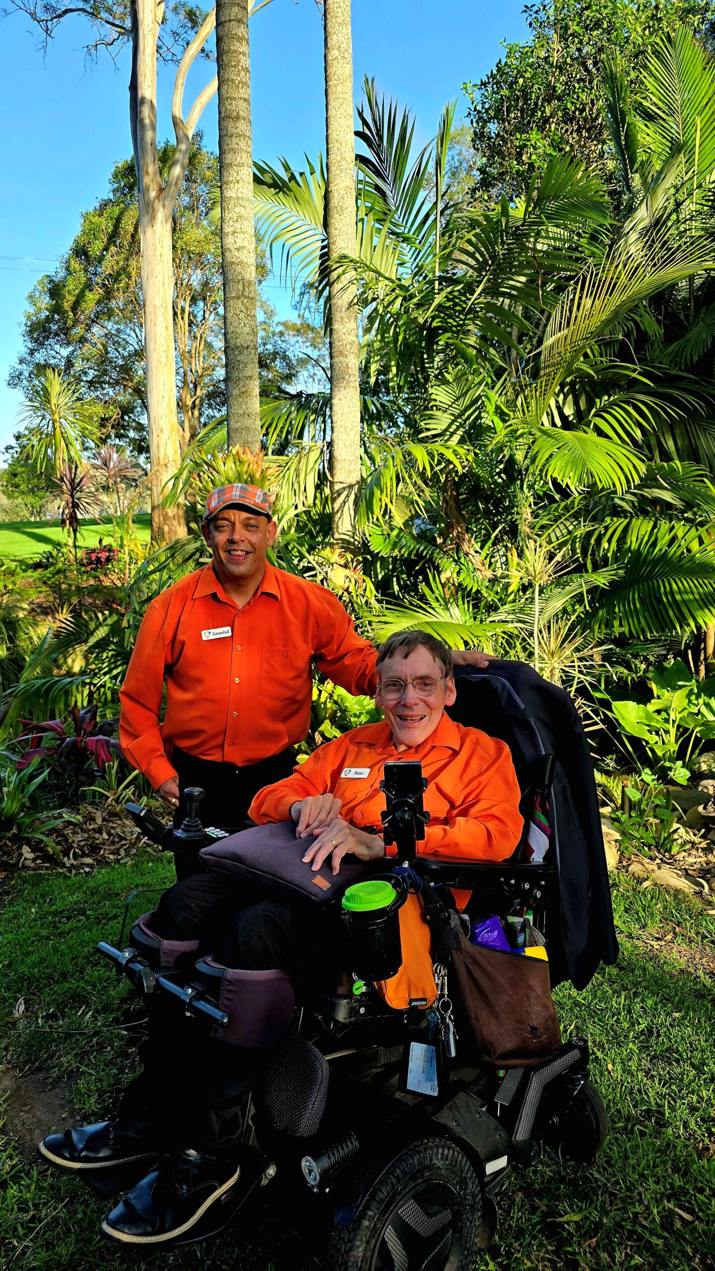 20251112_165517 Co-founders of DCAMS , both wearing matching oragne shirts, stand and sit in a tropical outdoor setting. Peter is in a power wheelchair, while Grandall stands beside him with a hand on peters shoulder. They are smiling.